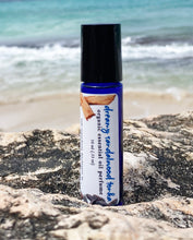 A 10 ml blue glass roller bottle, filled with essential oil perfume, rests on a rock at the beach with the ocean waves in the background