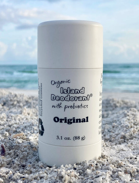 Island Deodorant Original Formula stick on white sand at the beach, ocean and sky in background. Natural, unscented deodorant with probiotics. Made in the USA.