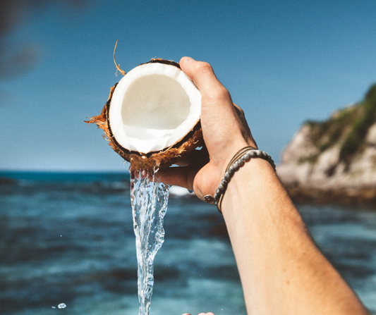 Hand holding fresh coconut pouring water with tropical ocean background – symbolizes natural ingredients like coconut oil in Island Deodorant’s Original Formula