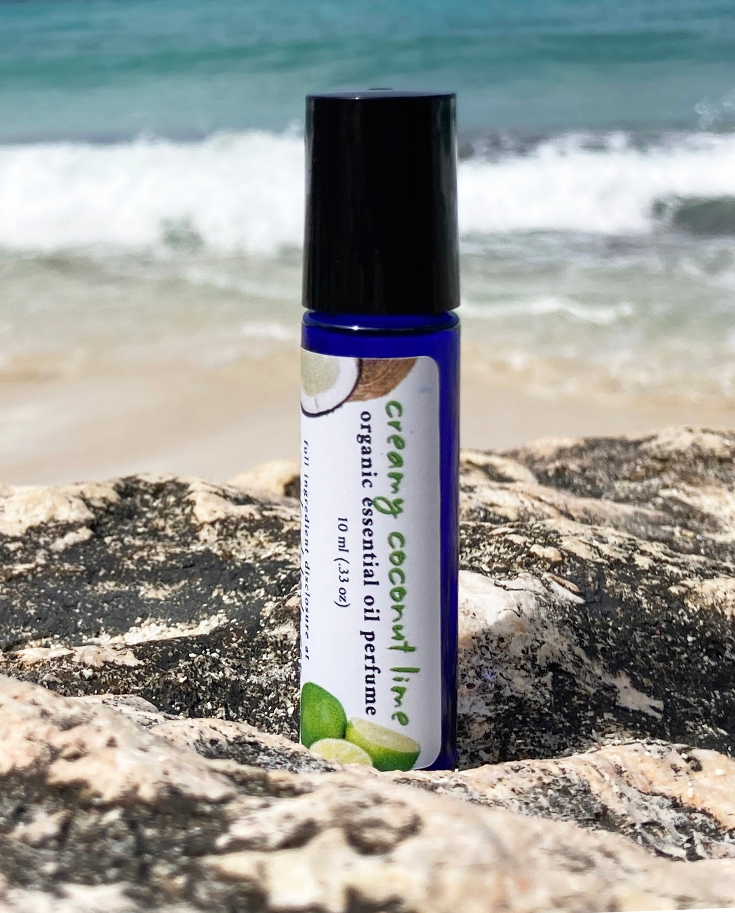 Coconut lime essential oil perfume roll-on bottle on a rocky beach with ocean waves in the background, representing a natural tropical scent.