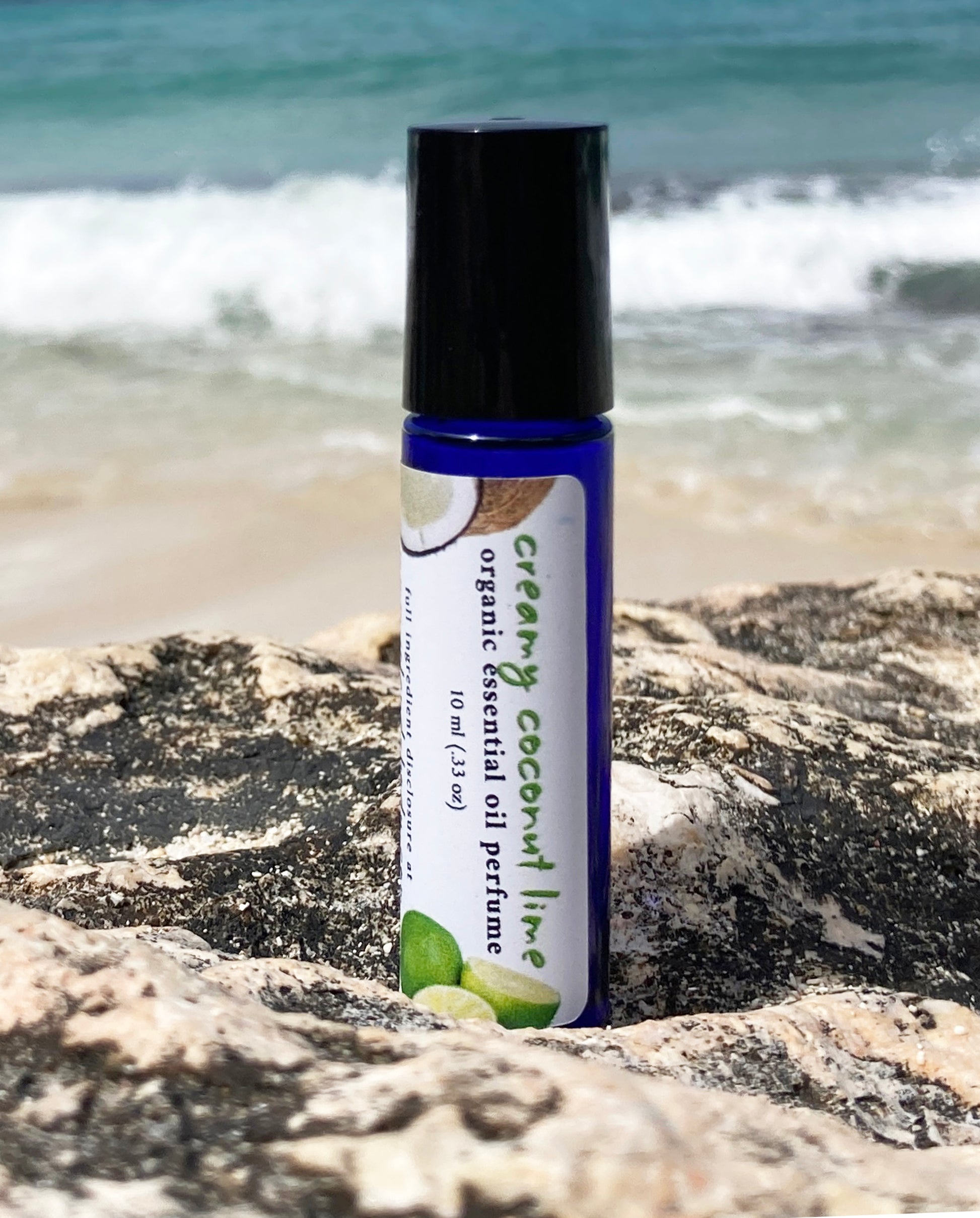 Coconut lime essential oil perfume roll-on bottle on a rocky beach with ocean waves in the background, representing a natural tropical scent.