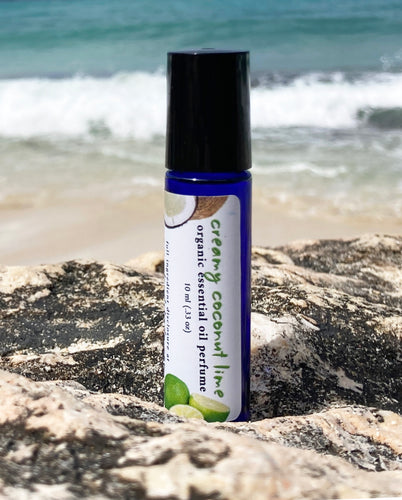 A 10 ml blue glass roller bottle, filled with essential oil perfume, rests on a rock at the beach with the ocean waves in the background