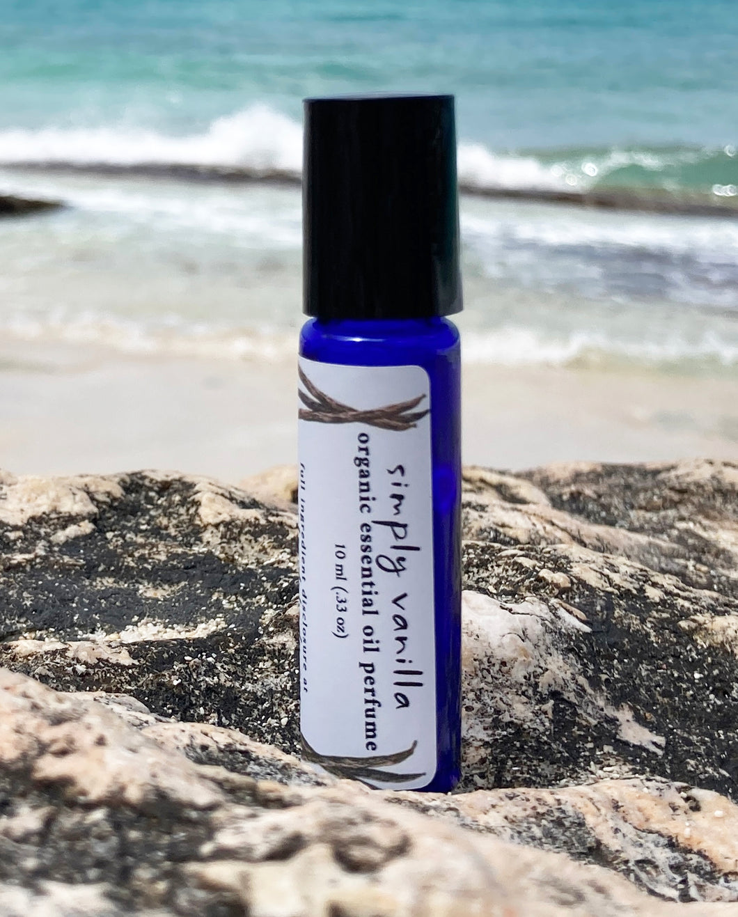 A 10 ml blue glass roller bottle, filled with essential oil perfume, rests on a rock at the beach with the ocean waves in the background
