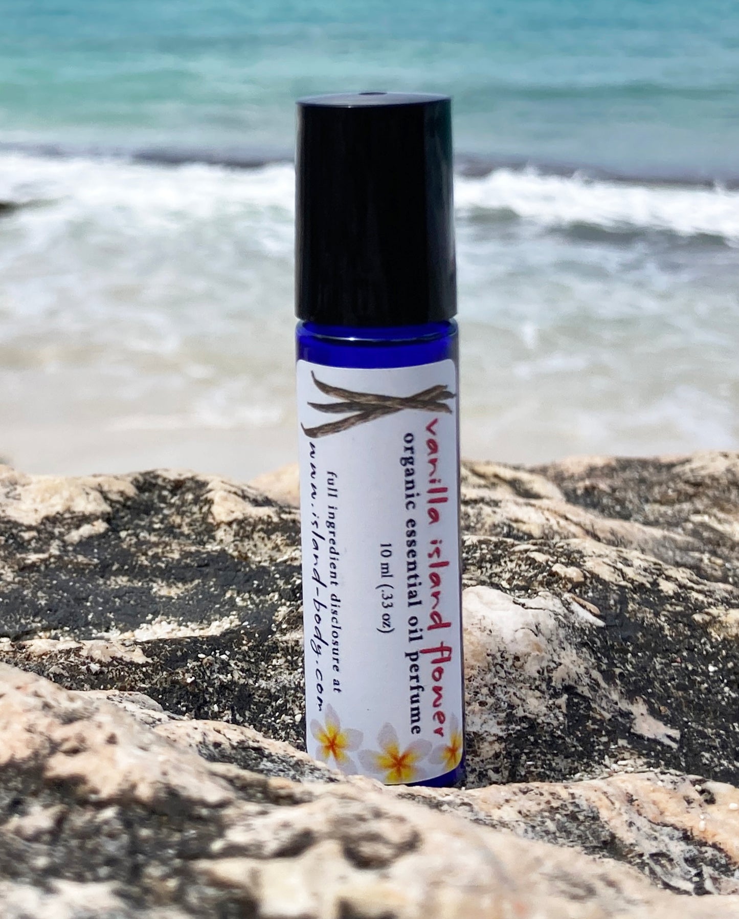 Vanilla Island Flower essential oil perfume roll-on bottle resting on a seaside rock with turquoise ocean waves in the background — natural tropical vanilla perfume by Island Deodorant