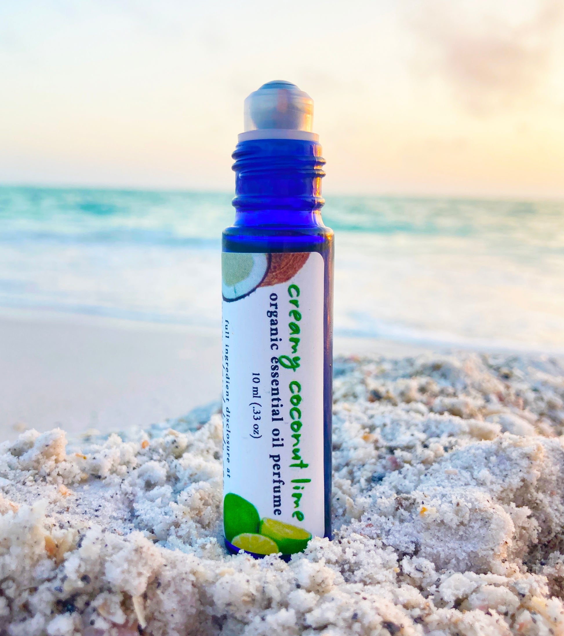 Blue glass roll-on bottle of Creamy Coconut Lime essential oil perfume resting in beach sand with the ocean and sunset in the background, evoking a natural tropical scent.